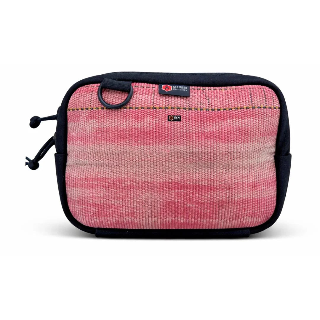 A Recycled Firefighter Trailhound fanny pack in Vintage Red firehose material. The fabric shows a weathered, authentic distress from years of service, featuring a matte red finish with a black zipper, black adjustable strap, and a side brass grommet for dog bags.